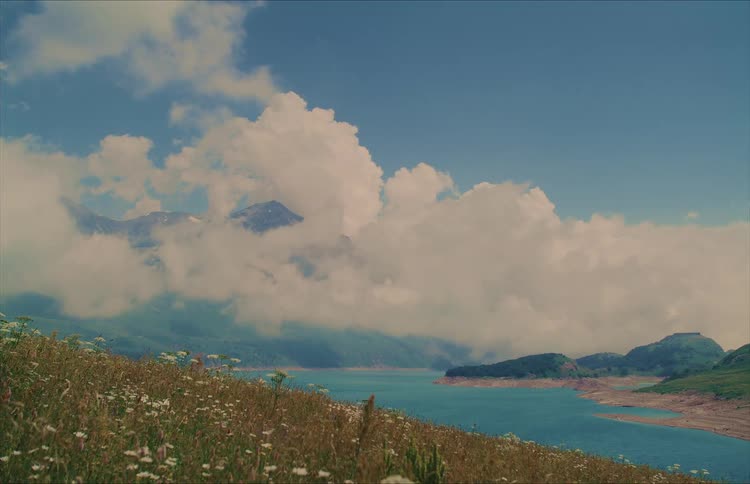 A stunning landscape of a tranquil mountain lake with fluffy white clouds covering the peaks and a field of wildflowers on the grassy hillside