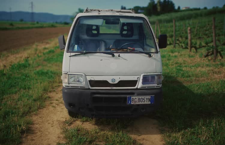 A static shot of an old white utility van parked on a dirt road in the Italian countryside next to a vineyard