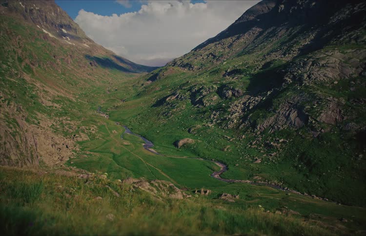 A serene view of a vast green mountain valley with a winding river flowing through it under a partly cloudy sky