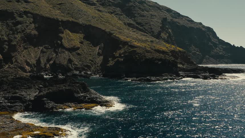 A scenic view of a rugged, mossy cliffside meeting the sparkling blue ocean on a sunny day