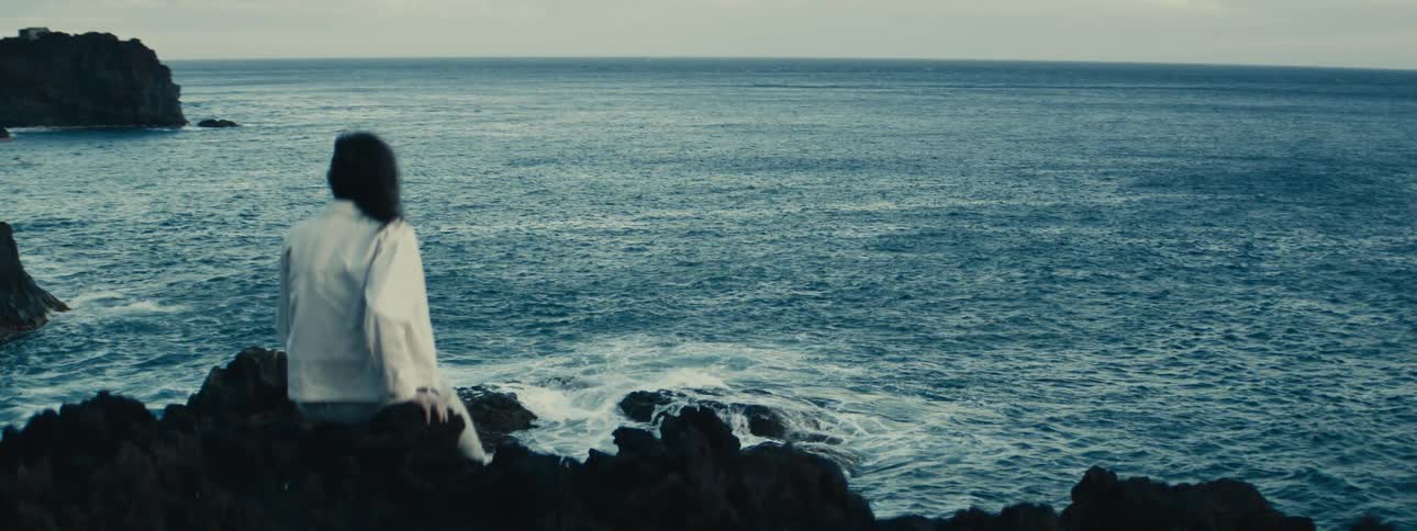 A lone figure sits on a dark, rocky coastline, contemplatively gazing out at the vast, rippling ocean