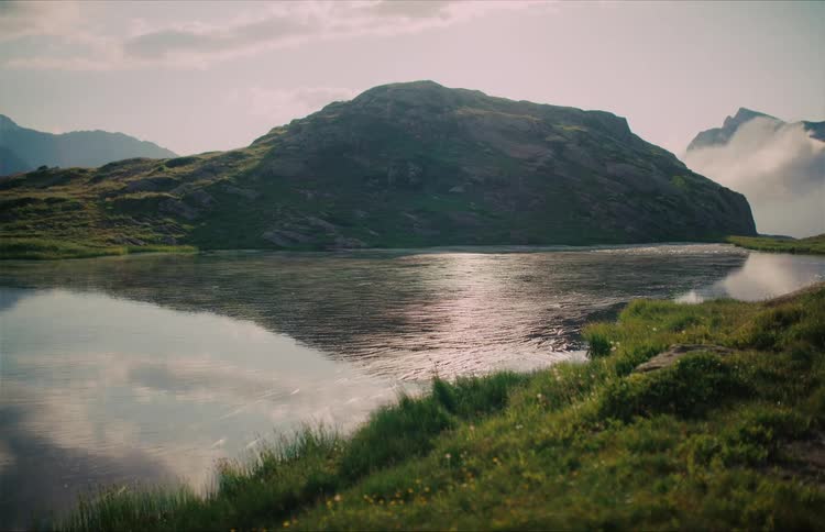 A tranquil alpine lake perfectly reflects the grassy mountain and cloudy sky on a calm day