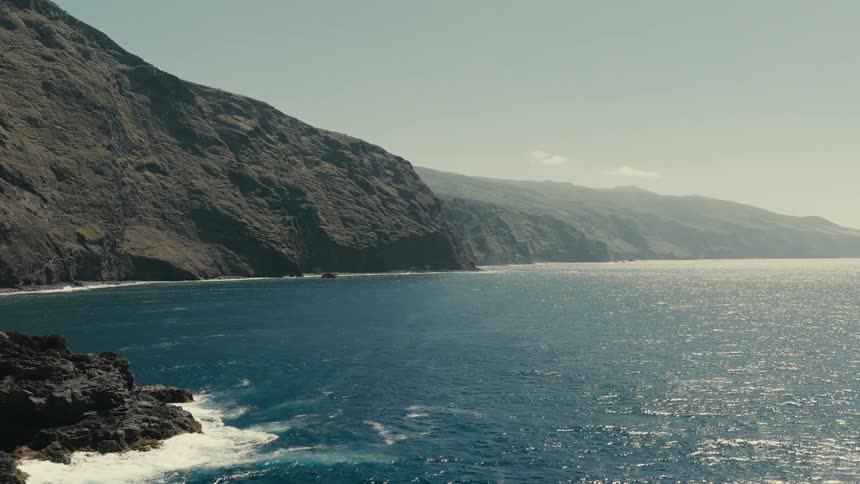 A serene view of a rugged, mountainous coastline meeting the sparkling blue ocean on a sunny day
