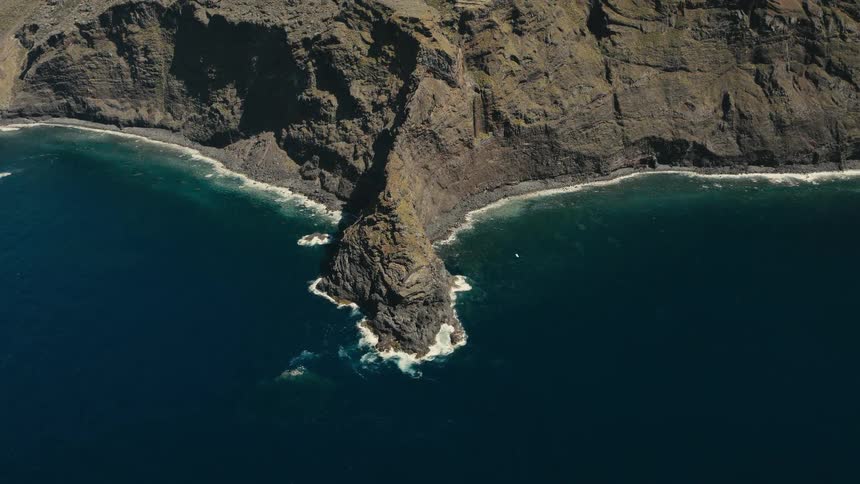 A static top-down aerial shot captures a rugged volcanic cliff meeting the deep blue ocean, with waves crashing around a prominent sea stack