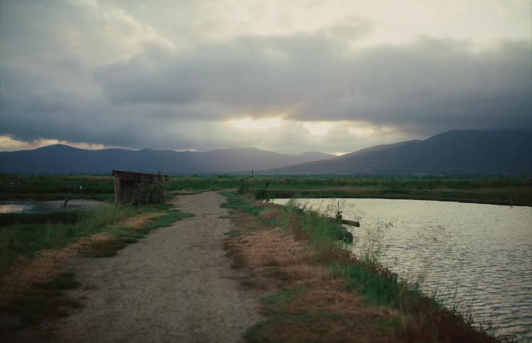 A tranquil dirt path through a rural marshland leads towards distant mountains under a cloudy, moody sunset