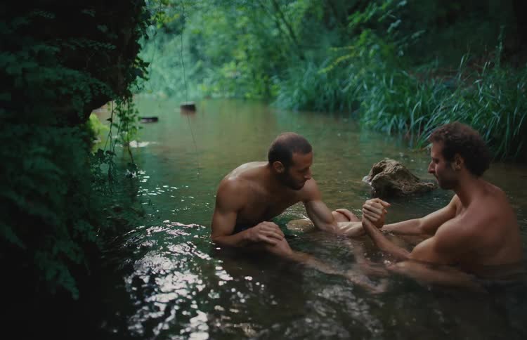 Two men arm wrestle while sitting in a serene river surrounded by a lush green forest