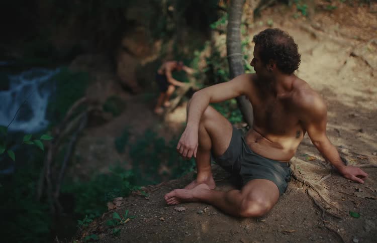 A shirtless man sits contemplatively on a dirt ledge in a lush forest, with a flowing waterfall visible in the background