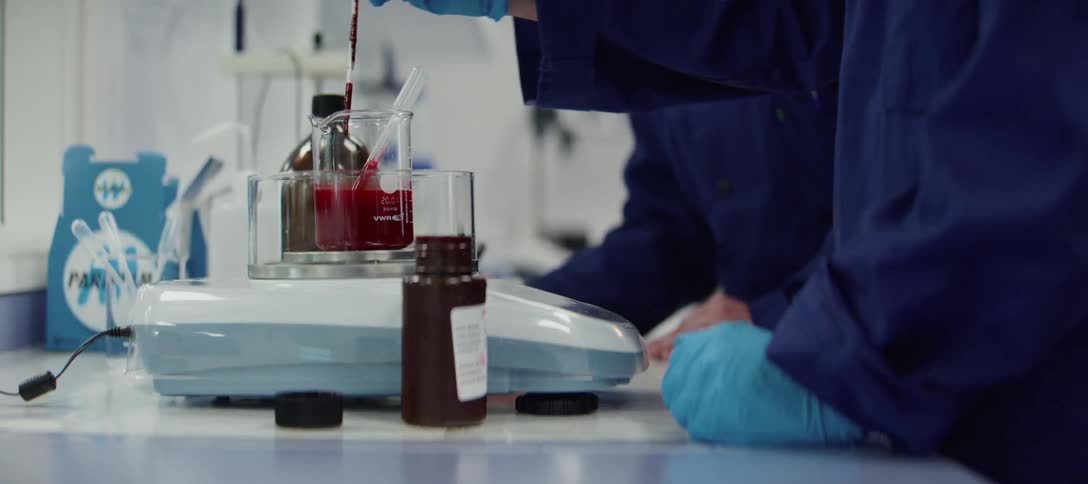 A gloved scientist carefully transfers a red liquid with a pipette into a beaker in a modern laboratory