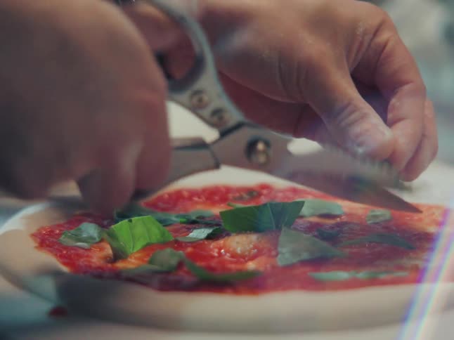 Close up shot of a chef's hands cutting fresh basil with scissors over a pizza