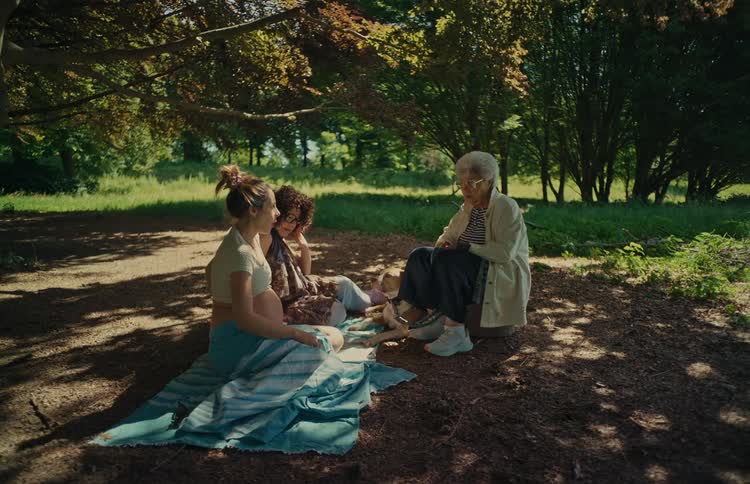 An elderly woman shares her wisdom with a pregnant woman and her friend during a relaxing picnic in a sunlit park