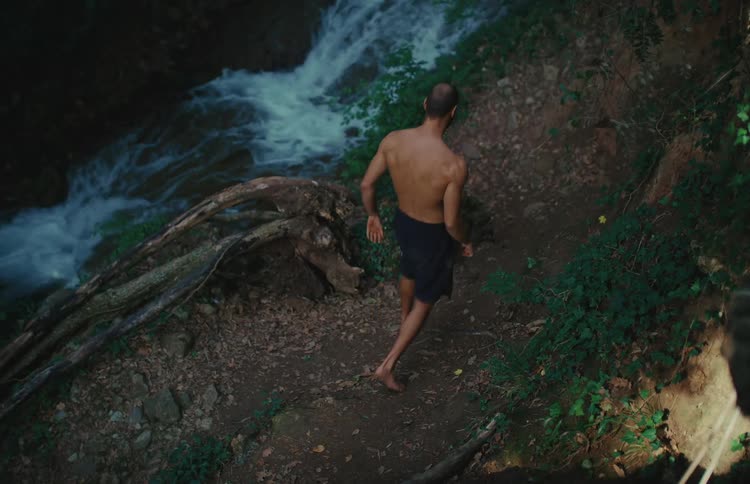 High-angle view of a shirtless man warming up on a forest path next to a rushing waterfall
