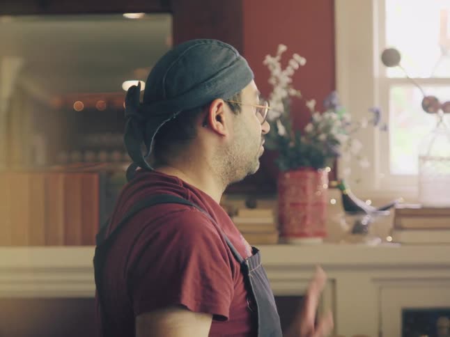 A side profile shot shows a man in a bandana and apron having a serious conversation while gesturing in a sunlit room