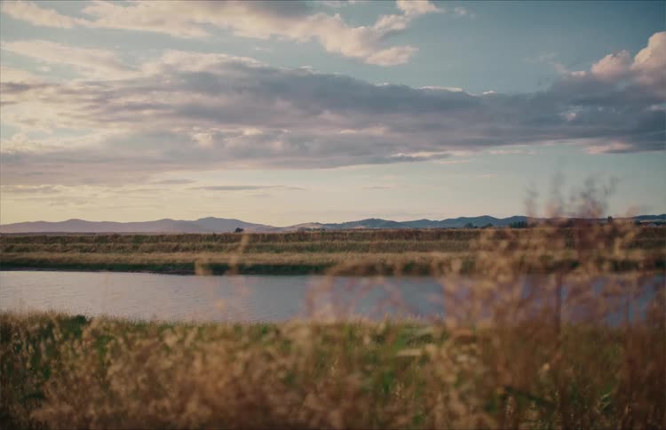 A serene wide shot of a peaceful river landscape with distant hills under a cloudy sky during the golden hour
