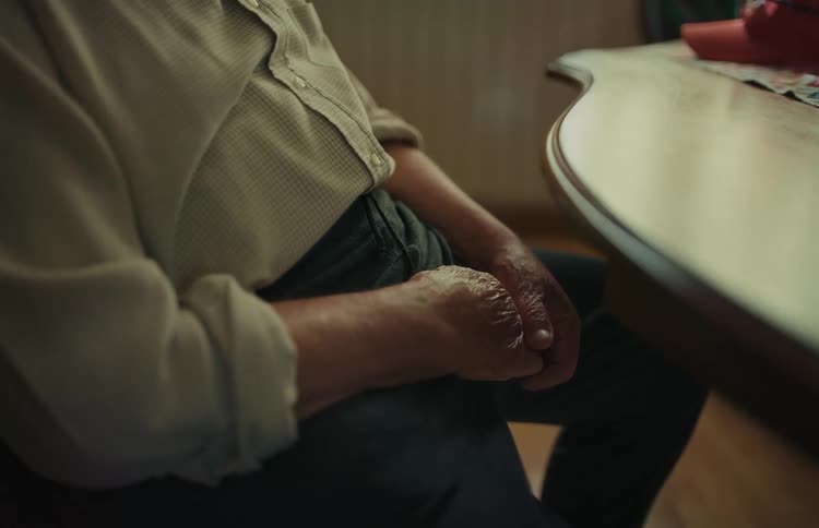 An elderly man rests his clasped, wrinkled hands on his lap while sitting alone at a table
