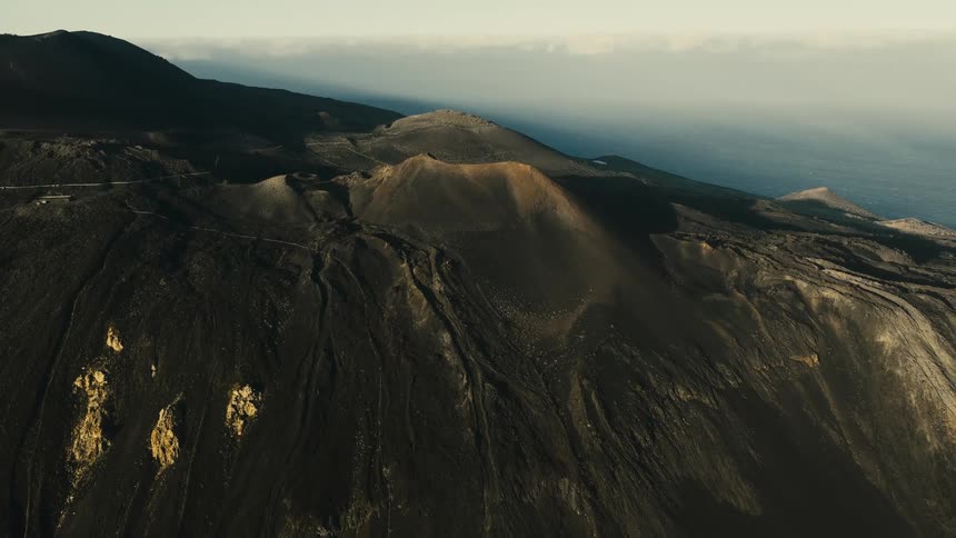 A stunning aerial view pulls back to reveal the dramatic, dark volcanic craters and landscape of a coastal mountain at sunrise