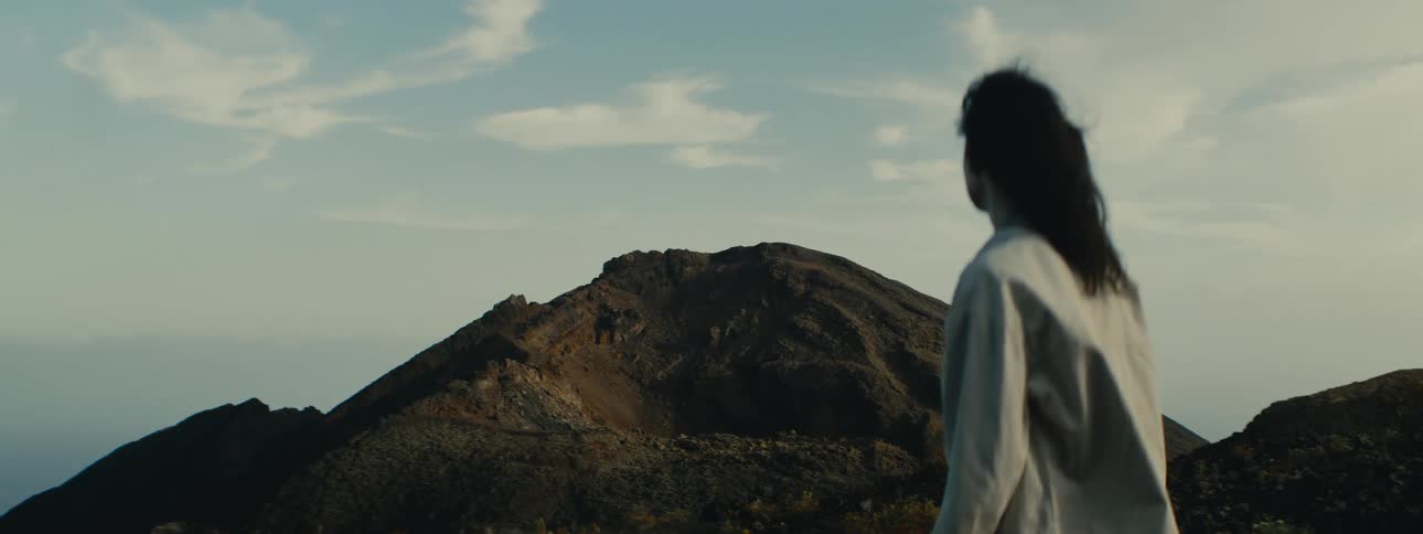 An over-the-shoulder shot captures a person with dark, flowing hair looking out at a magnificent dormant volcano