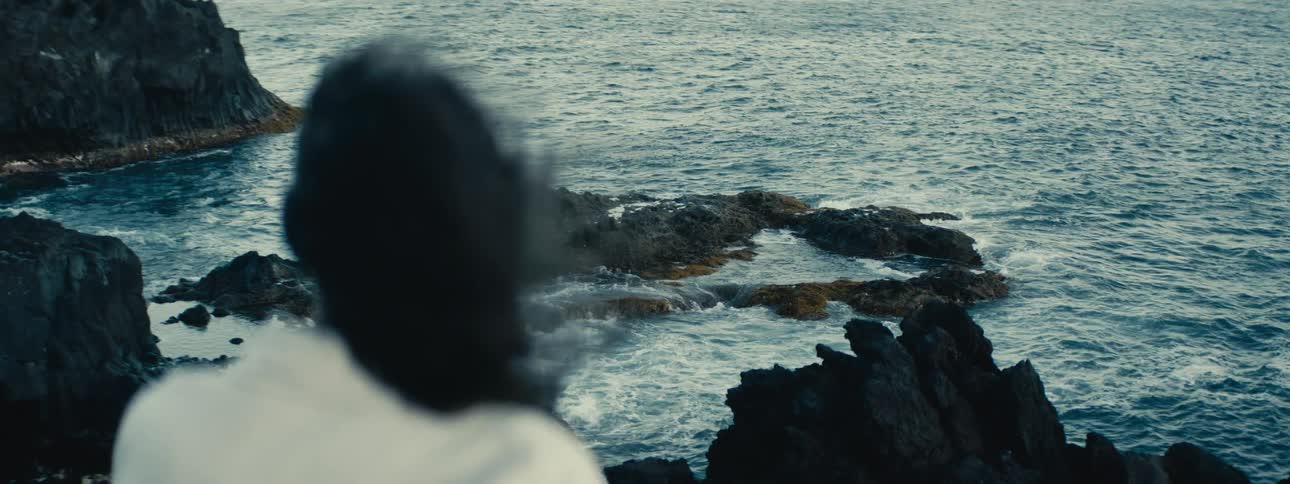 A person with hair blowing in the wind contemplates the dramatic ocean waves crashing against a rocky coastline