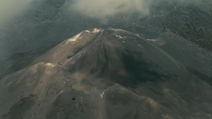 An aerial view descends through thick clouds, slowly revealing the barren, ashen peak and craters of a massive volcano