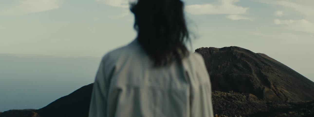 A solitary figure with long hair blowing in the wind stands looking out over a vast and desolate volcanic mountain landscape