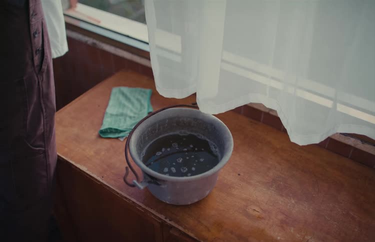 A metal bucket with soapy water sits on a wooden surface next to a window with a sheer curtain blowing in the wind