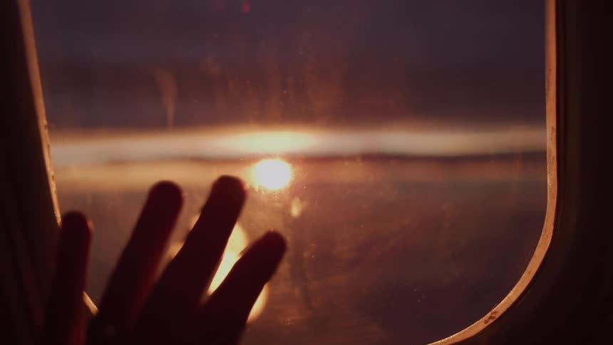 A dreamy, slow-motion shot pulls focus from a beautiful sunset over the clouds to a person's hand resting on the airplane window