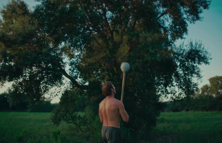A shirtless muscular man performs a slow-motion workout with a steel mace in a serene green field at sunset