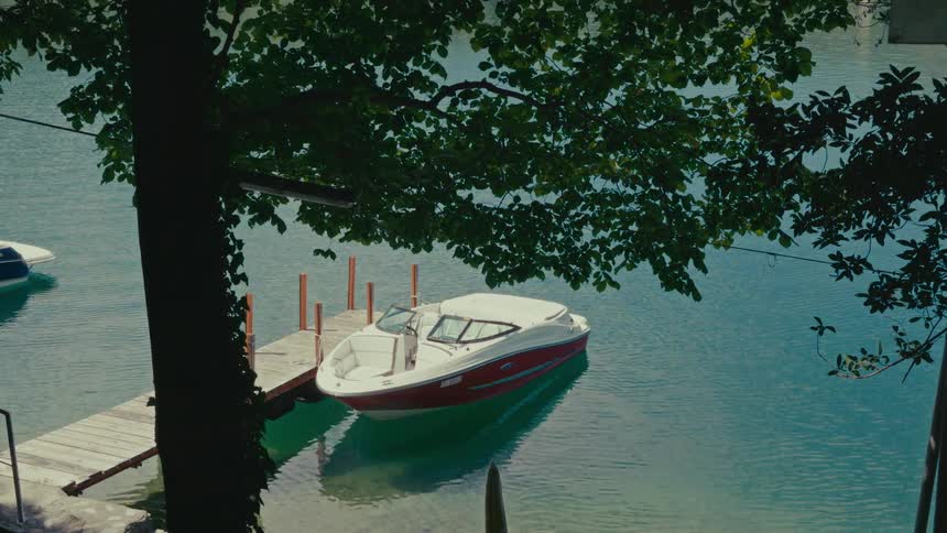 A red and white motorboat is docked at a wooden pier on a calm, turquoise lake, viewed from behind a leafy tree