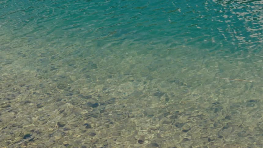 Sunlight reflects off the gentle ripples of crystal clear turquoise water, revealing a rocky bottom
