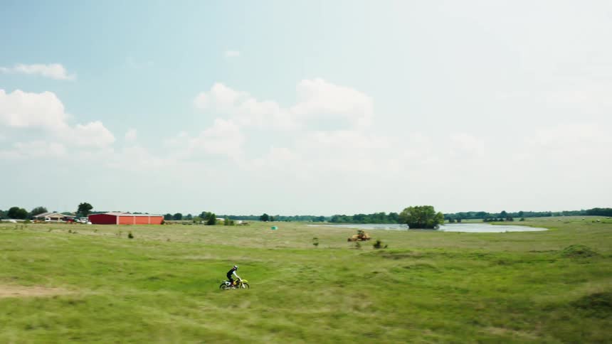 A motocross rider speeds along a winding dirt track through a sprawling green field under a bright blue sky