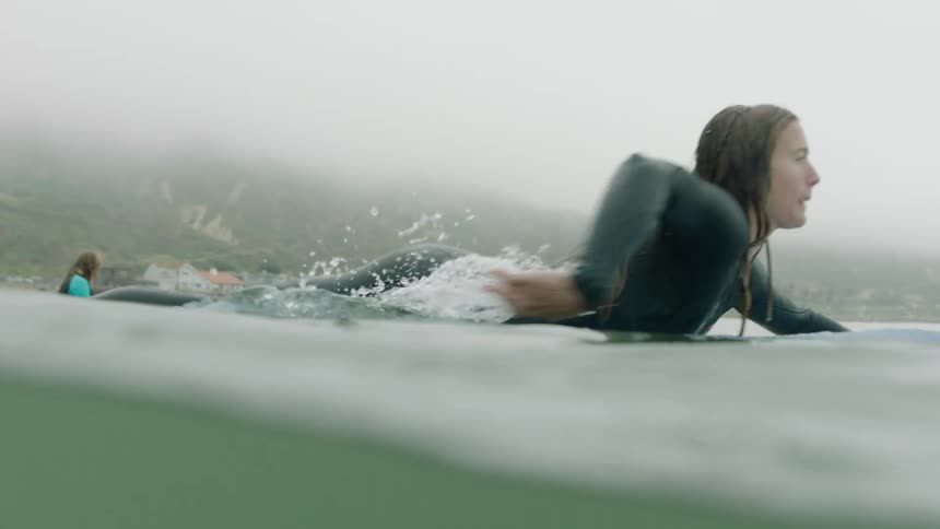 A female surfer in a wetsuit paddles her surfboard through the calm, foggy ocean waters in slow motion