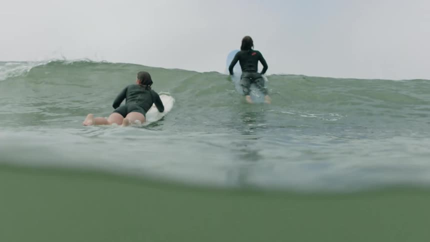 From a water-level view in slow motion, two female surfers in wetsuits paddle on their boards over the crest of a wave