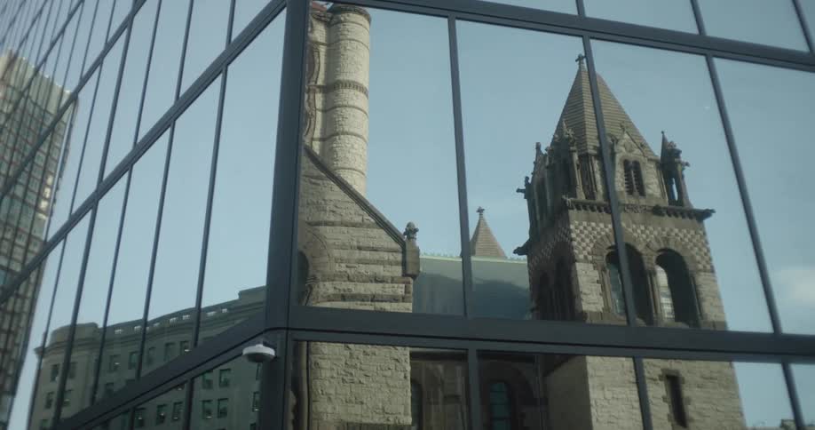 A slow camera move reveals the reflection of a historic stone church and other city buildings in the glass facade of a modern skyscraper