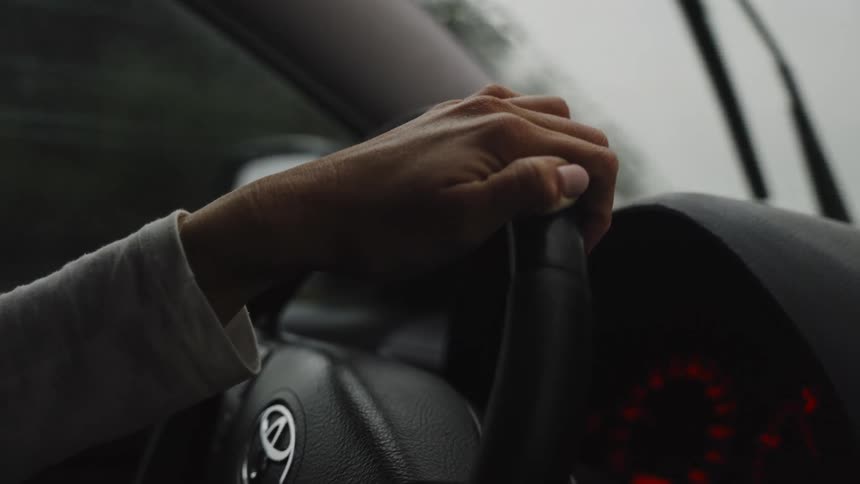 A close-up of a person's hand holding the steering wheel while driving on a rainy day
