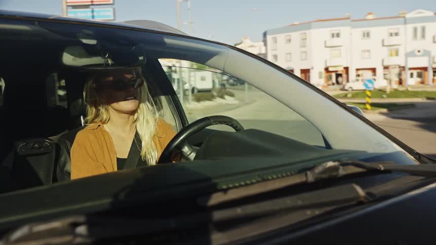 A young blonde woman wearing sunglasses drives her car through a town on a sunny day
