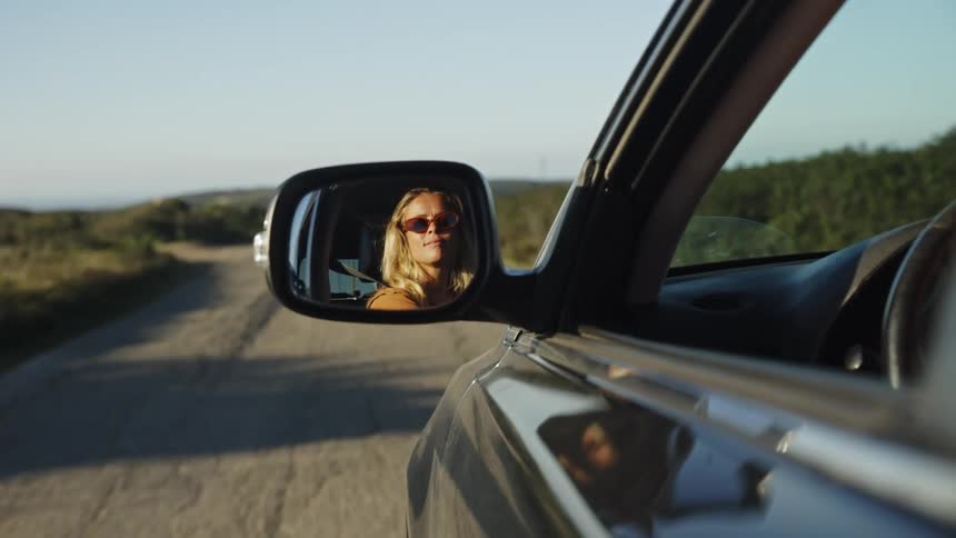 Stylish blonde woman wearing sunglasses is reflected in the car's side-view mirror while driving down a scenic country road