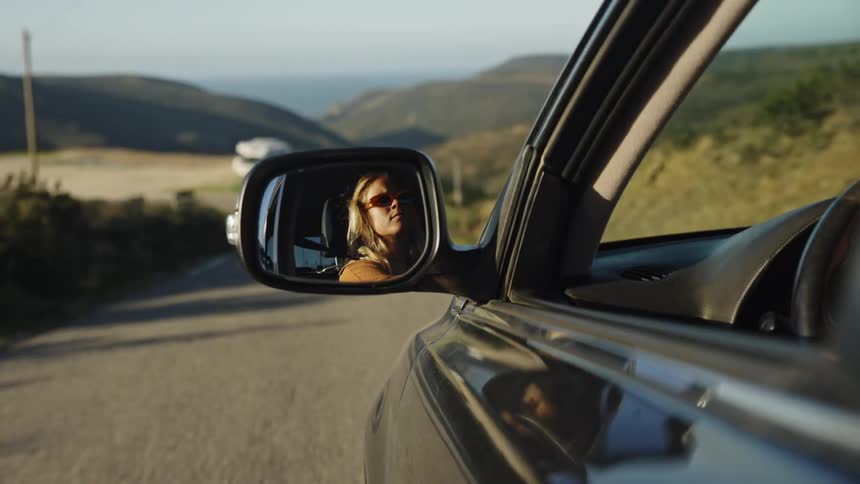 Reflection of a woman with sunglasses in a car's side-view mirror while driving on a scenic road trip at golden hour