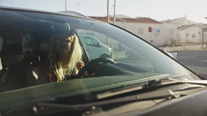 A stylish blonde woman in sunglasses drives her car in reverse through a sun-drenched parking lot