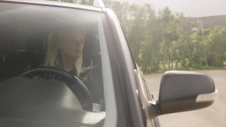 A young blonde woman is seen through the windshield of her car as she drives along a scenic country road