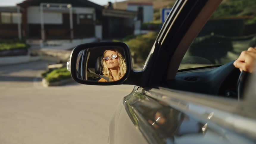 A blonde woman wearing sunglasses is reflected in the side-view mirror of a car during a scenic drive at golden hour