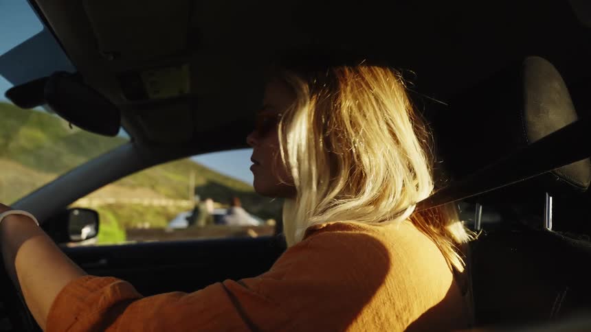 A stylish blonde woman with sunglasses drives a car along a scenic route as the golden hour sunlight streams through the window