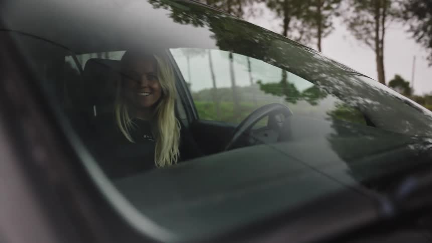 A smiling young blonde woman drives a car down a road, seen through the windshield in slow motion