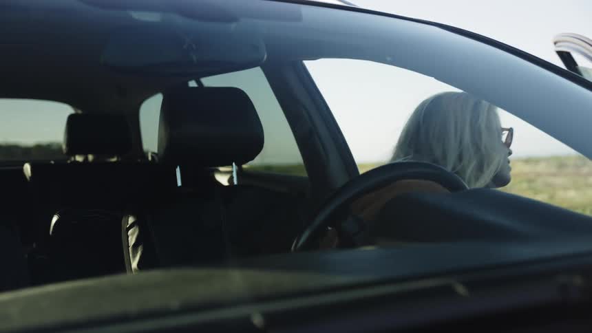 A young blonde woman in sunglasses finishes her drive, parks on the side of the road, and exits her car on a sunny day