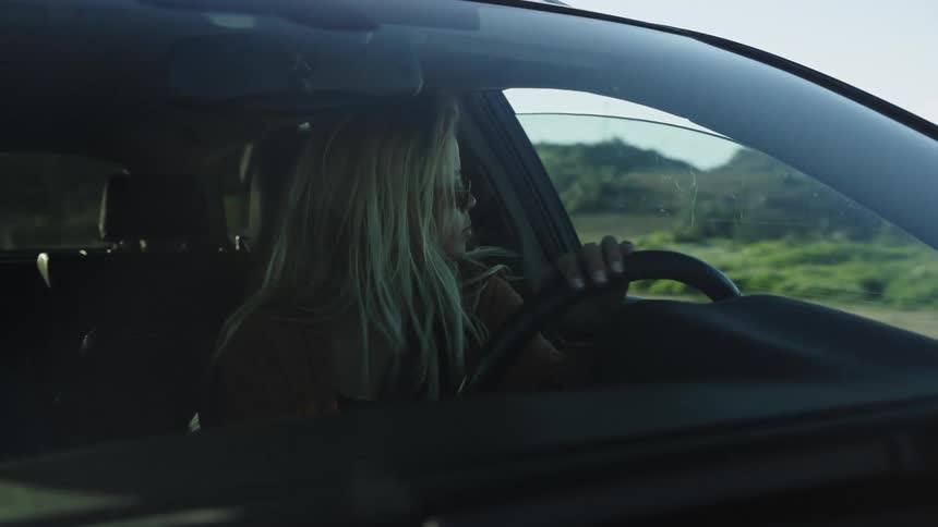 A stylish blonde woman wearing sunglasses drives her car along a scenic route with the sun flaring through the windshield