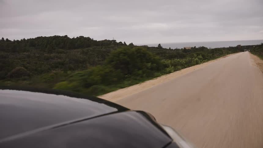 POV from a black car driving along a dusty coastal road with the ocean in the distance on a cloudy day
