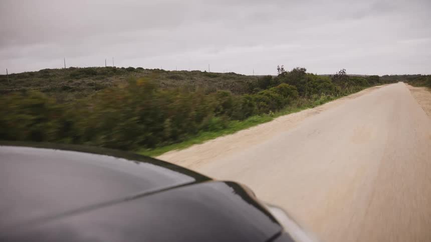 A point-of-view shot from a car driving along a desolate, sandy dirt road through a rural landscape under an overcast sky