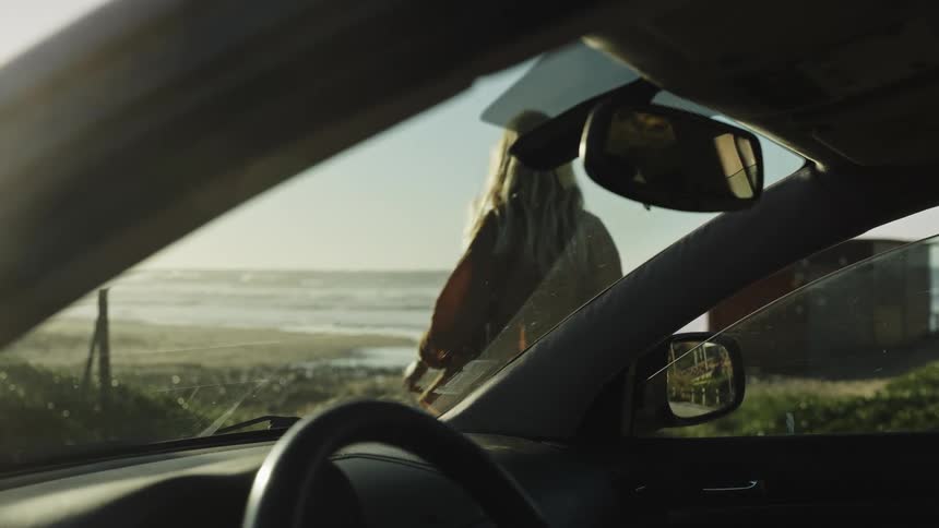 View from inside a car as a woman with a surfboard gets out and walks towards the ocean waves at sunrise