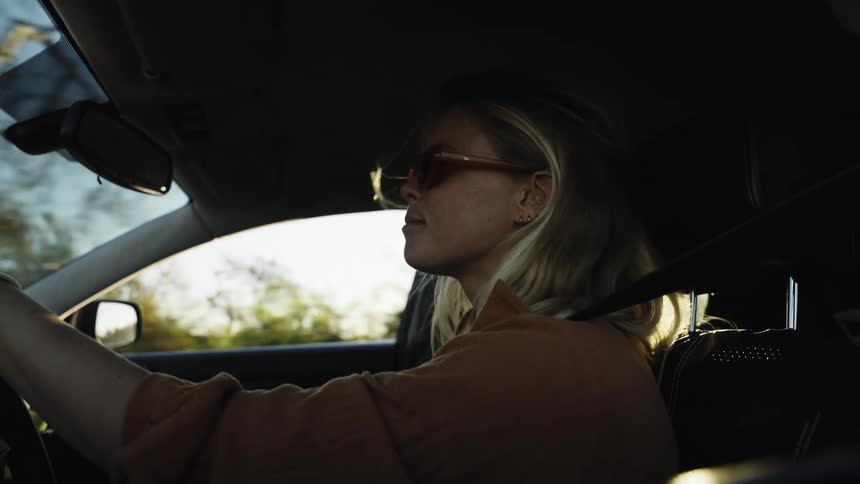 Slow motion close-up of a stylish young woman in sunglasses driving her car during a beautiful golden hour sunset
