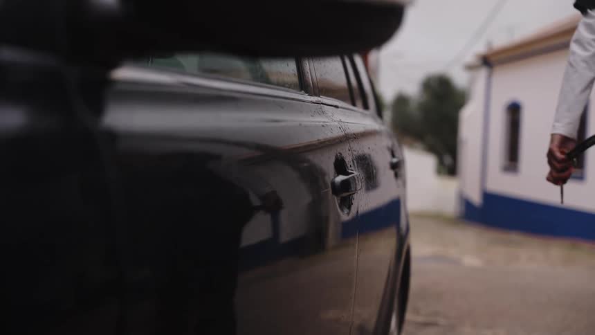 A person locks a black car with a key on a wet, overcast day, and their reflection is seen walking away with a dog