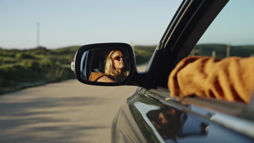 A carefree woman with sunglasses enjoys a scenic drive at golden hour, her reflection captured in the car's side mirror