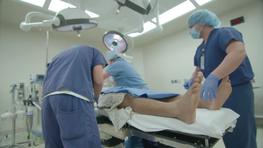 A medical team prepares a patient for surgery, starting with a reflex test on the foot in a brightly lit operating room